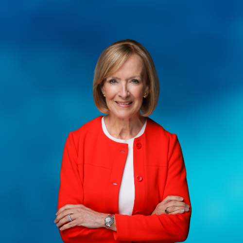 Judy Woodruff, arms crossed, in an orange jacket with a blue background.